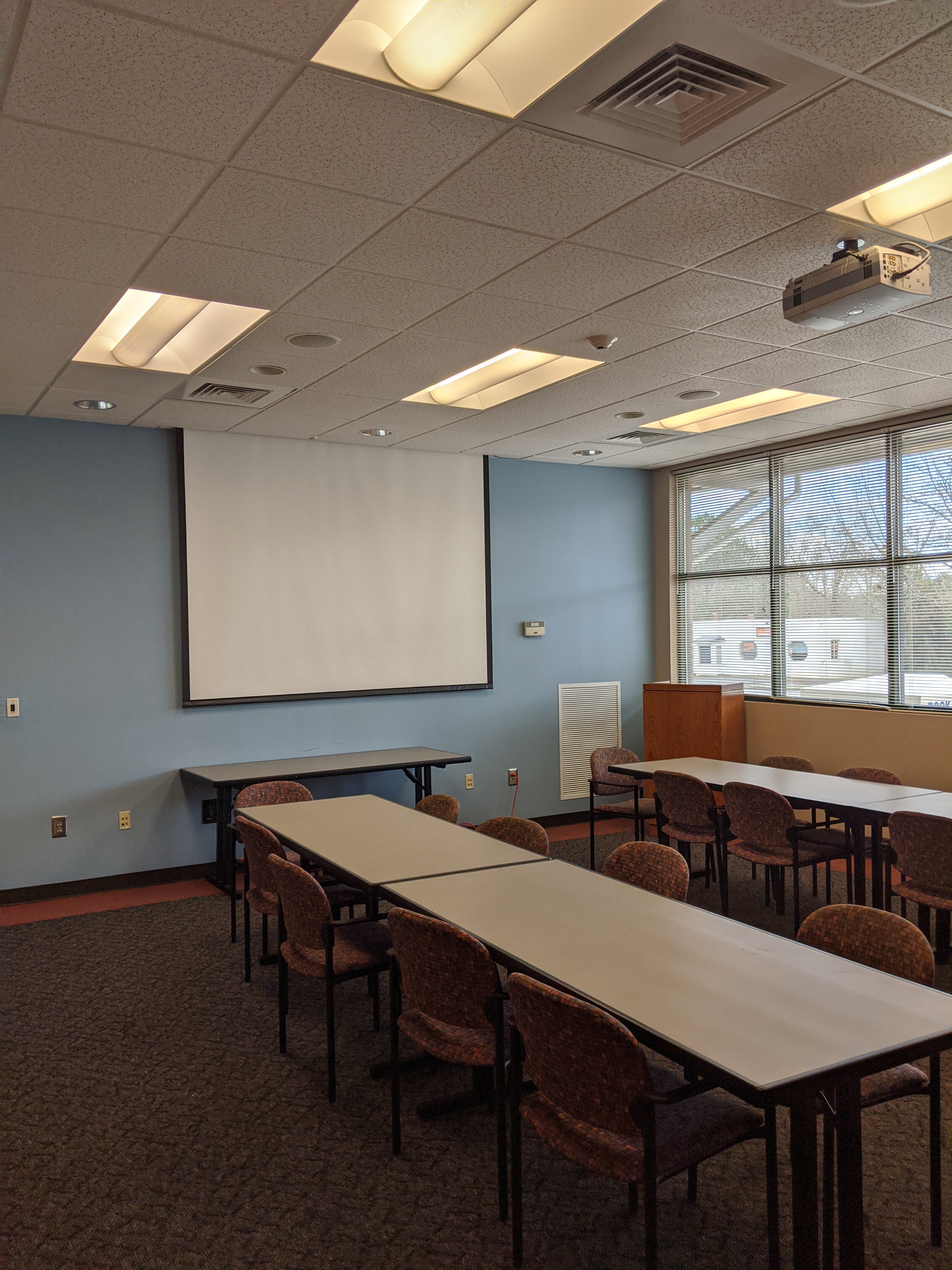 Room photo of the Friends of the Library Meeting Room showing tables placed parallel with a projector screen at the front of the room