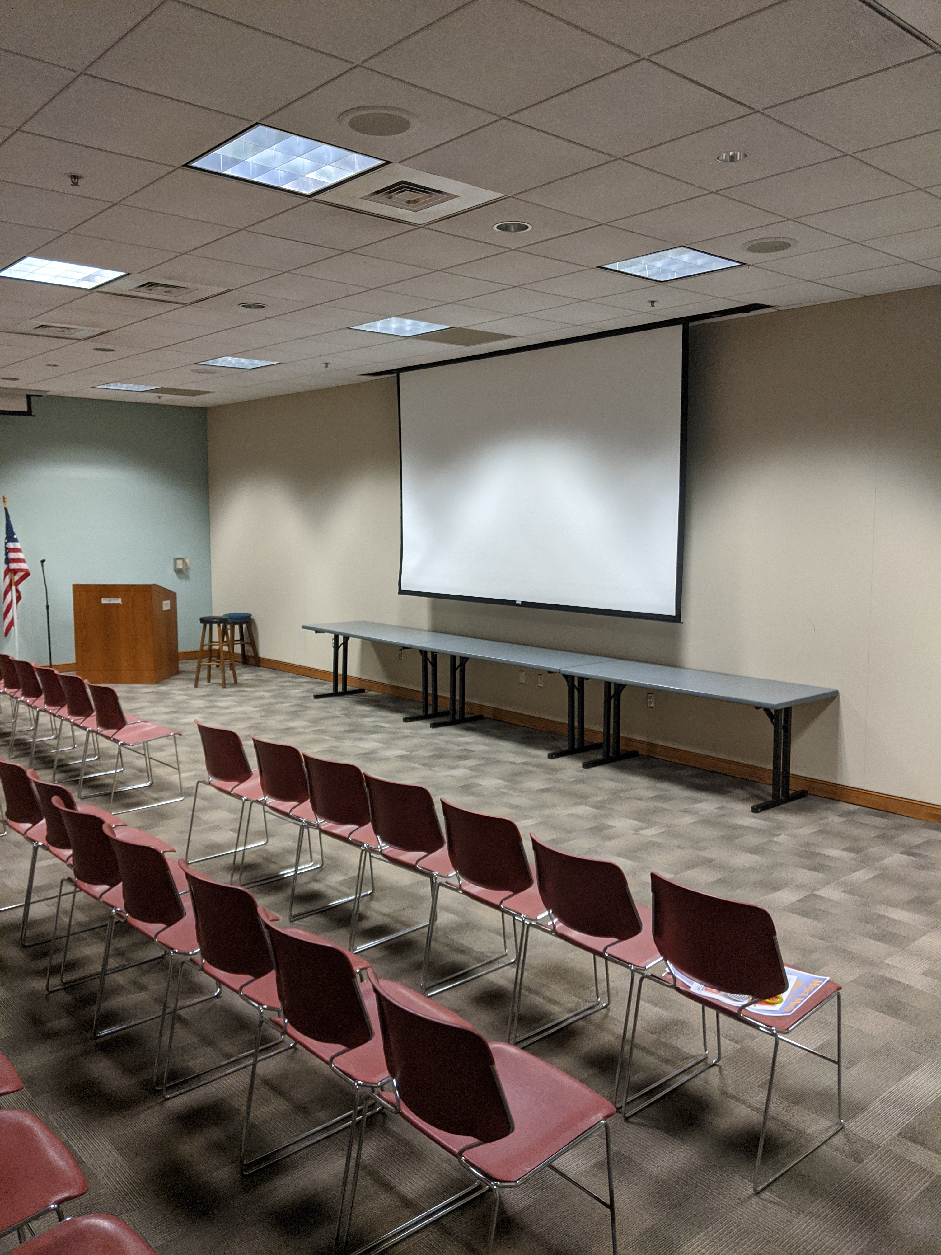 Room photo of Multipurpose Room C showing chairs placed in auditorium setup with projector screen pulled down at front