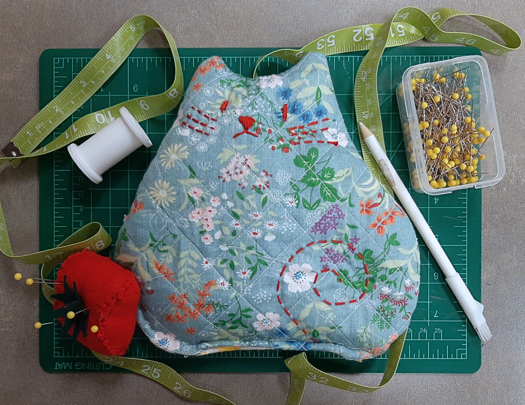 A cat shaped heating pad with embroidered details surrounded by sewing supplies 