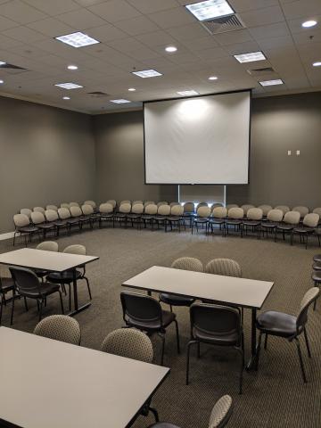 Cely Room photo showing tables, chairs, and extra chairs stacked on walls and projector screen